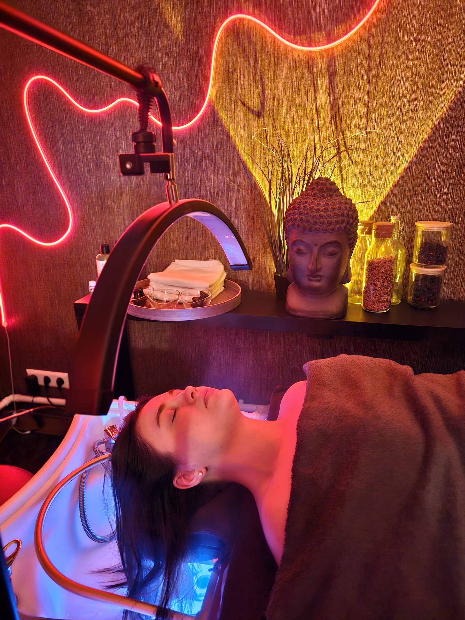 Een klant tijdens de headspa onder de lamp, met de boeddha en de kruidenpotten op de plank